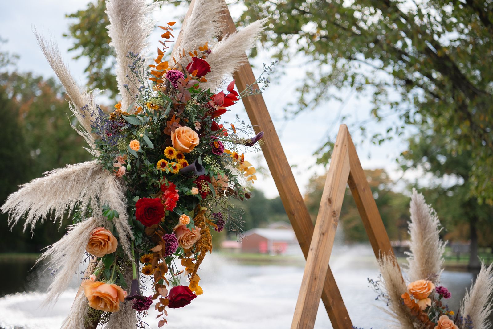 Triangle ceremony arch with pampas grass, burnt orange and red florals by the pond
